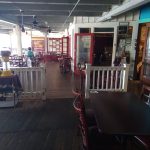 Open-air restaurant patio with wooden floor, dark chairs, and red-framed doorway leading inside.