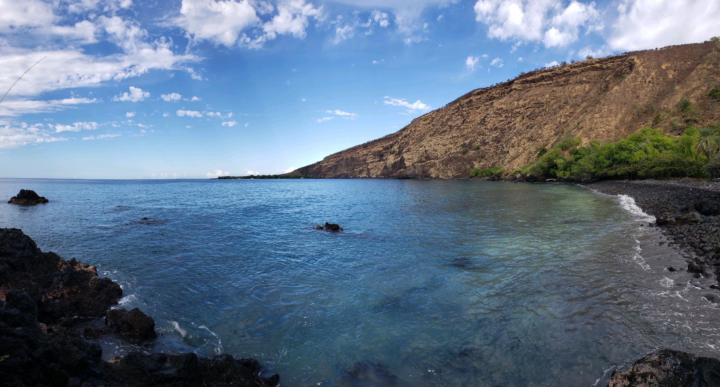 Blue sea along a rocky shore with a steep brown hillside covered in green trees on the right under a partly cloudy sky.