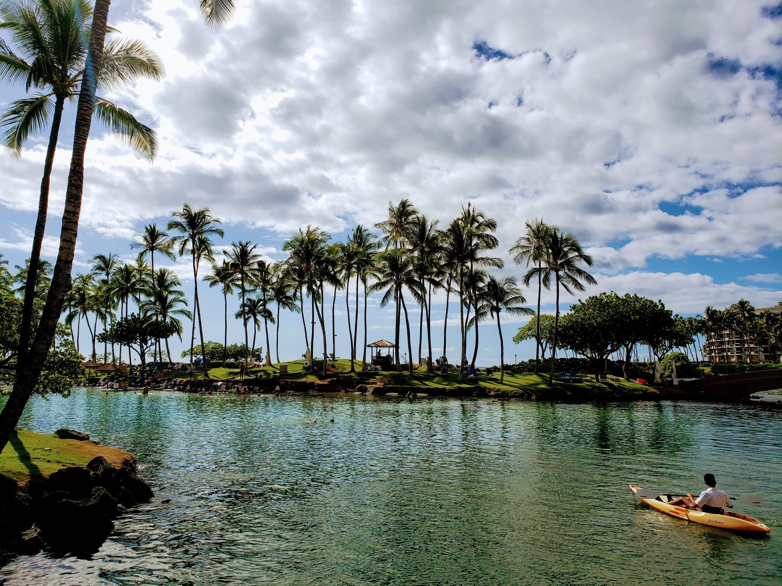 Palm trees line a lagoon where people relax on the grass and a small boat is on the water under a cloudy sky.