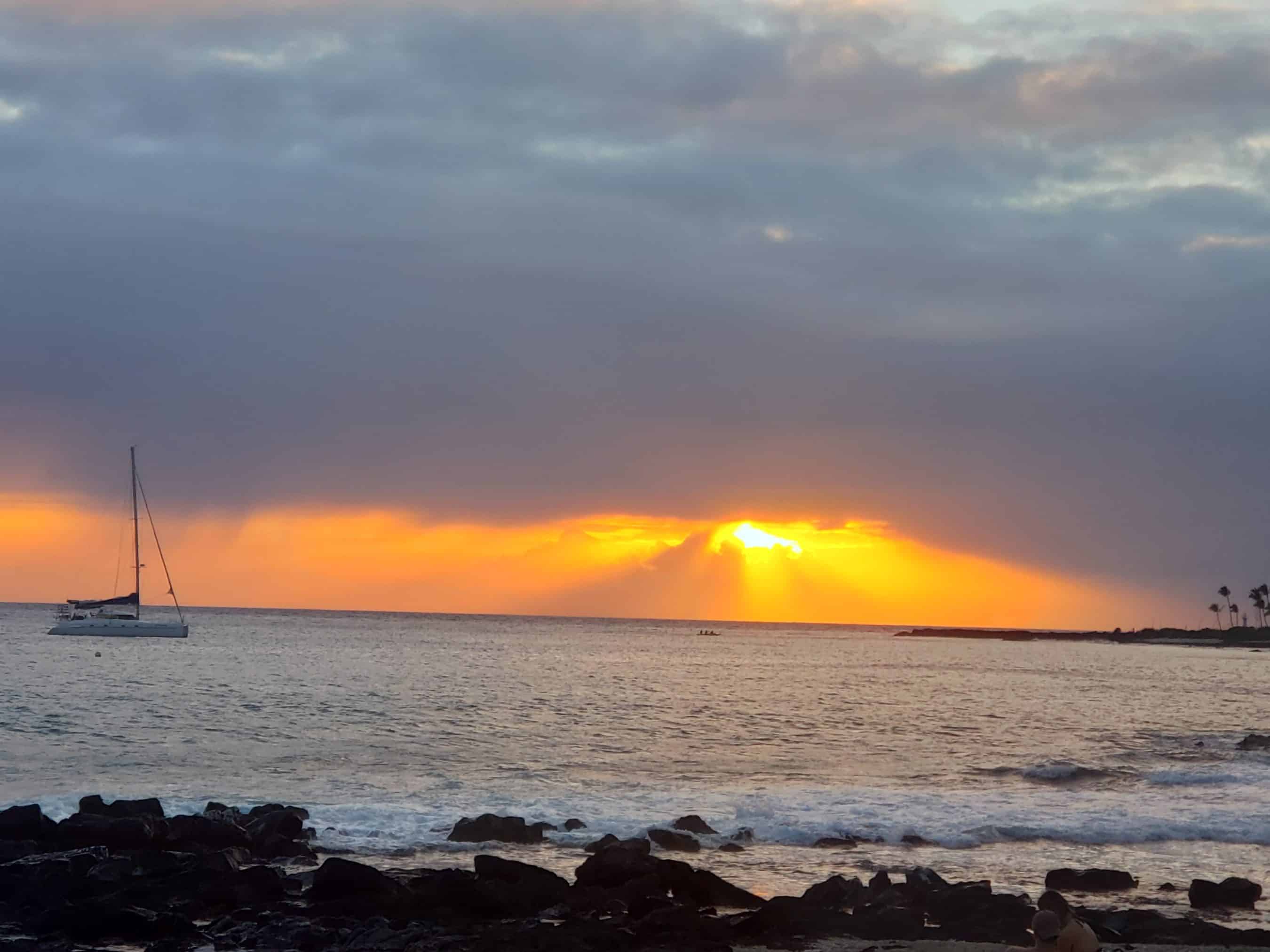 Sunset over the calm sea with an orange glow near the horizon, a small sailboat on the left, and dark rocks in the foreground.