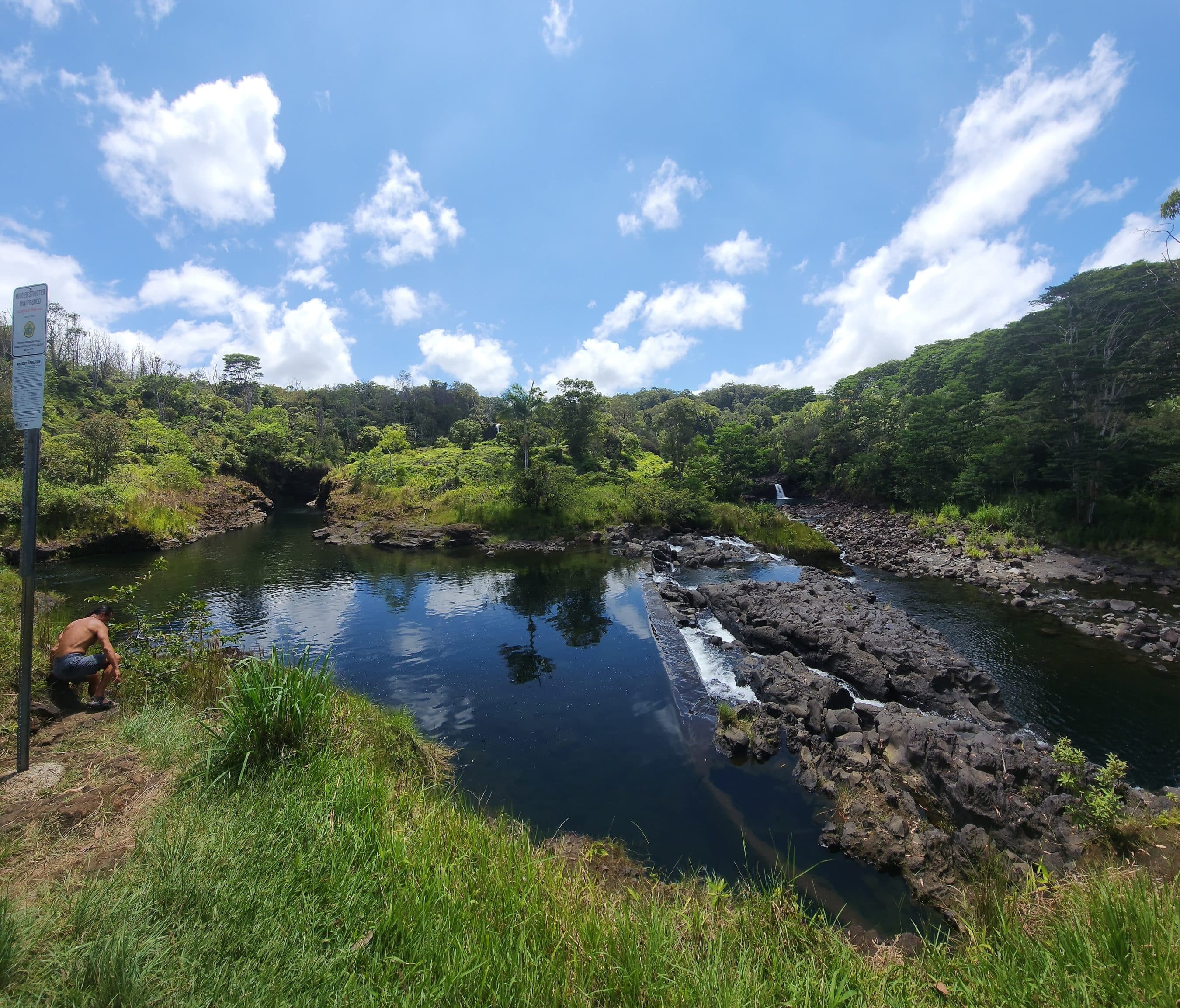 River with rocky banks and green vegetation, a signpost on the left, and a man crouching by the water at the edge; clear blue sky above.