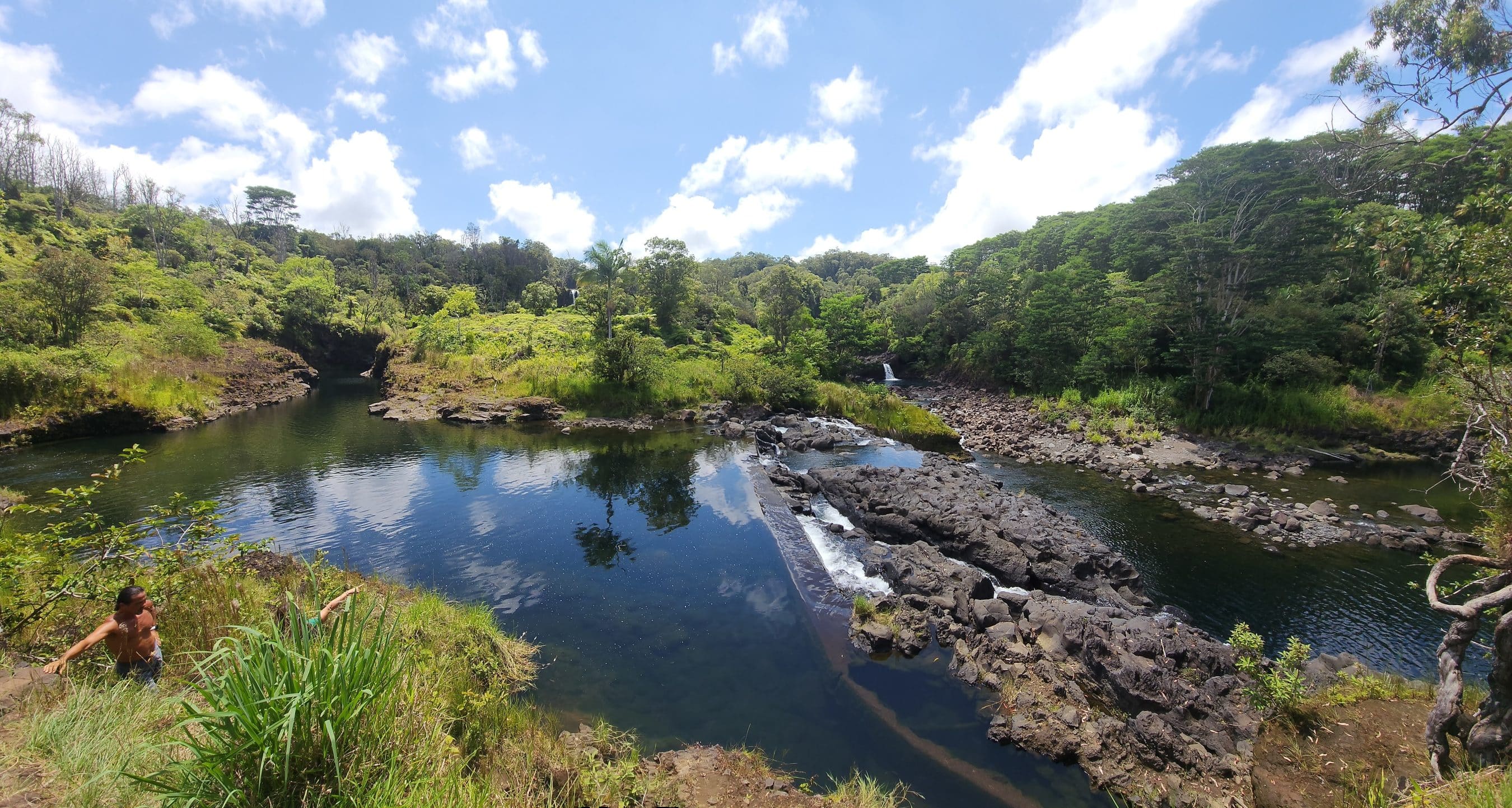 River winds through rocky banks with dense greenery and a bright blue sky; a shirtless person stands in the lower-left foreground.