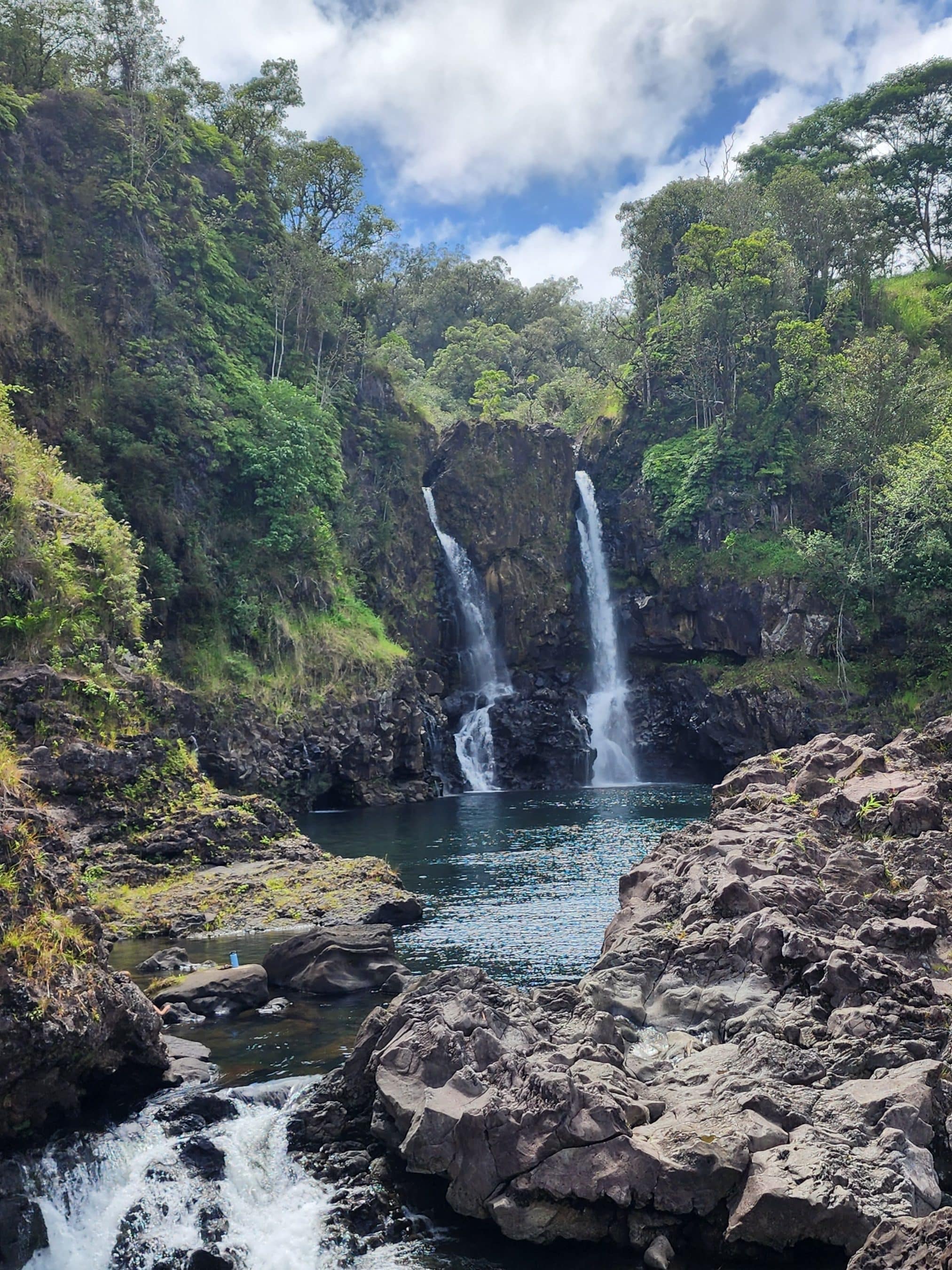 Twin waterfalls flow down a rocky gorge into a dark pool, surrounded by lush green trees and cliffs on a sunny day.