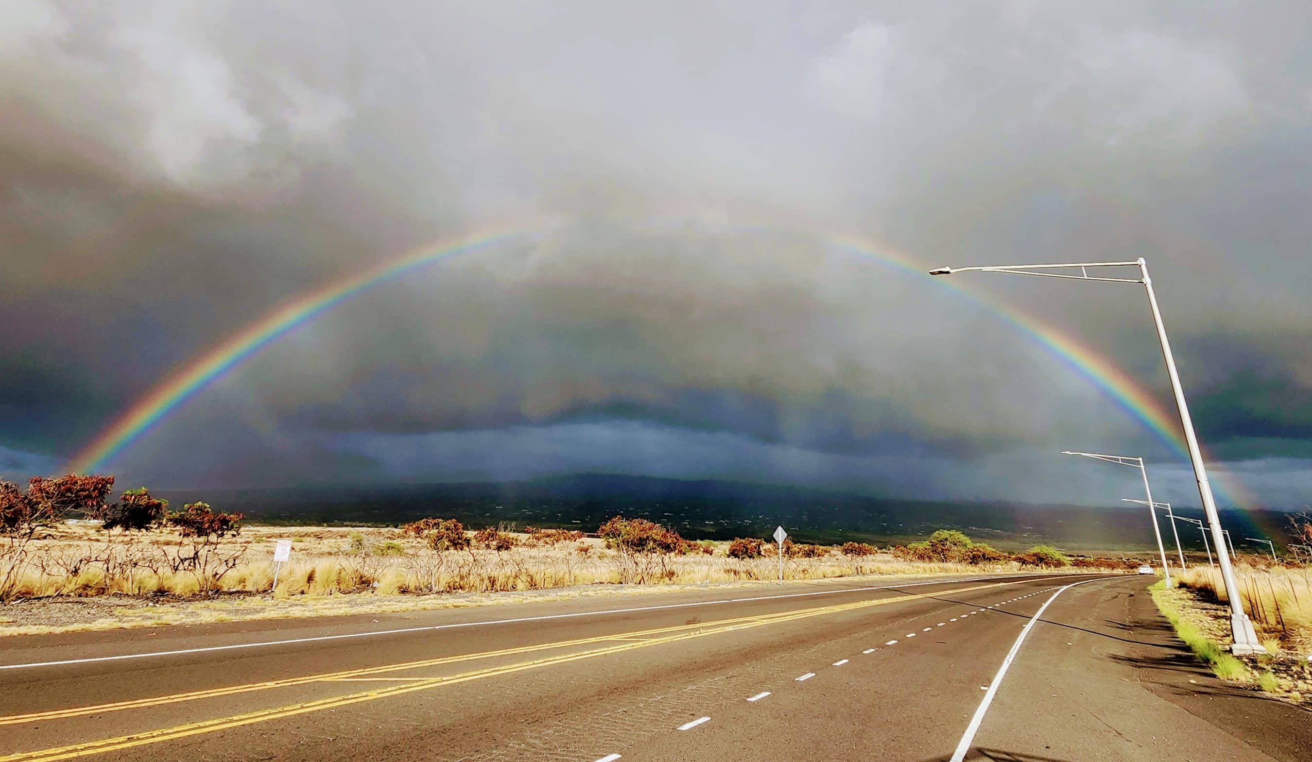 Double rainbow arching across a cloudy sky over a road and desert brush.