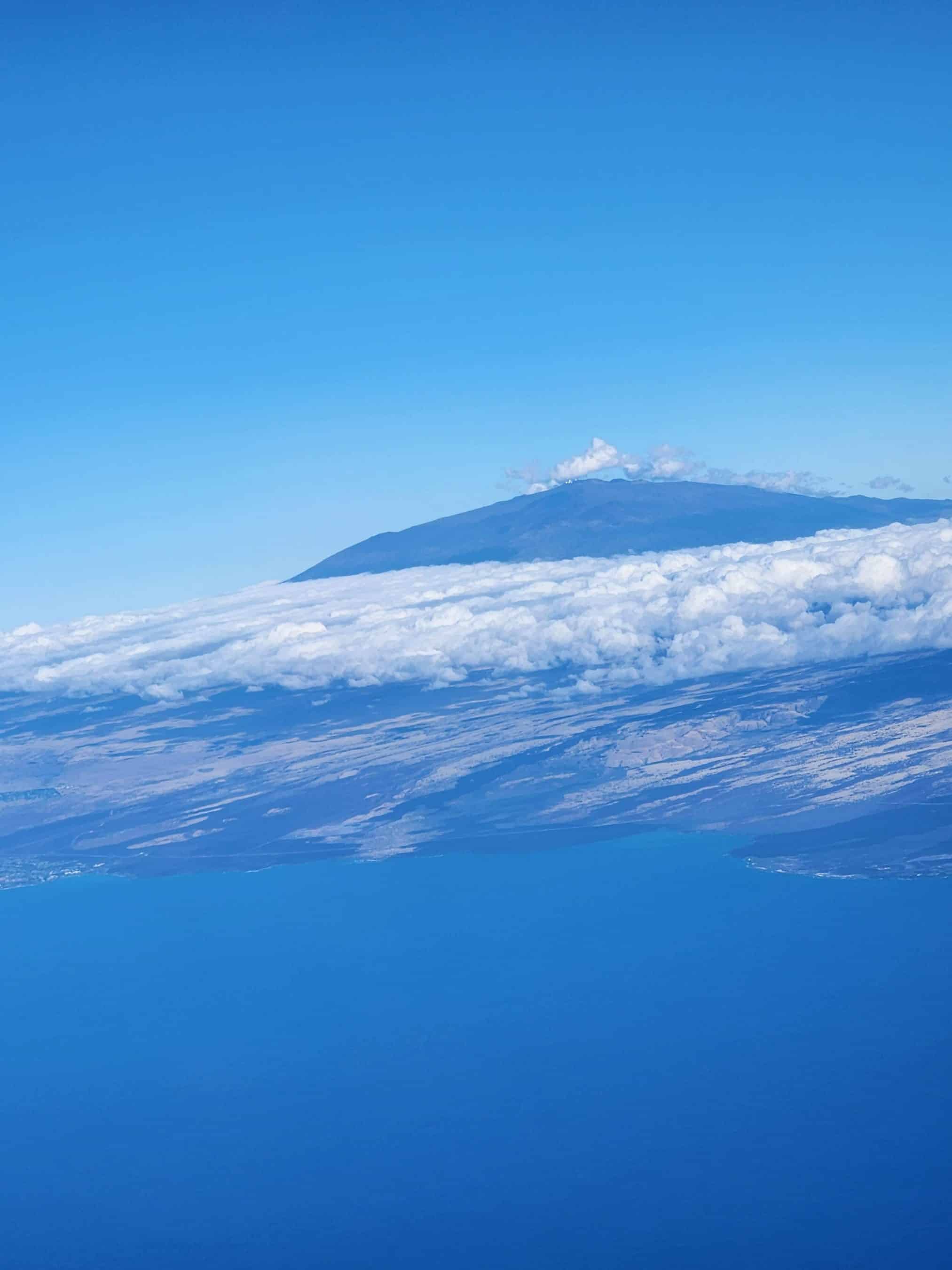 Snow-covered mountain peak rising above a blanket of white clouds under a clear blue sky (aerial view).