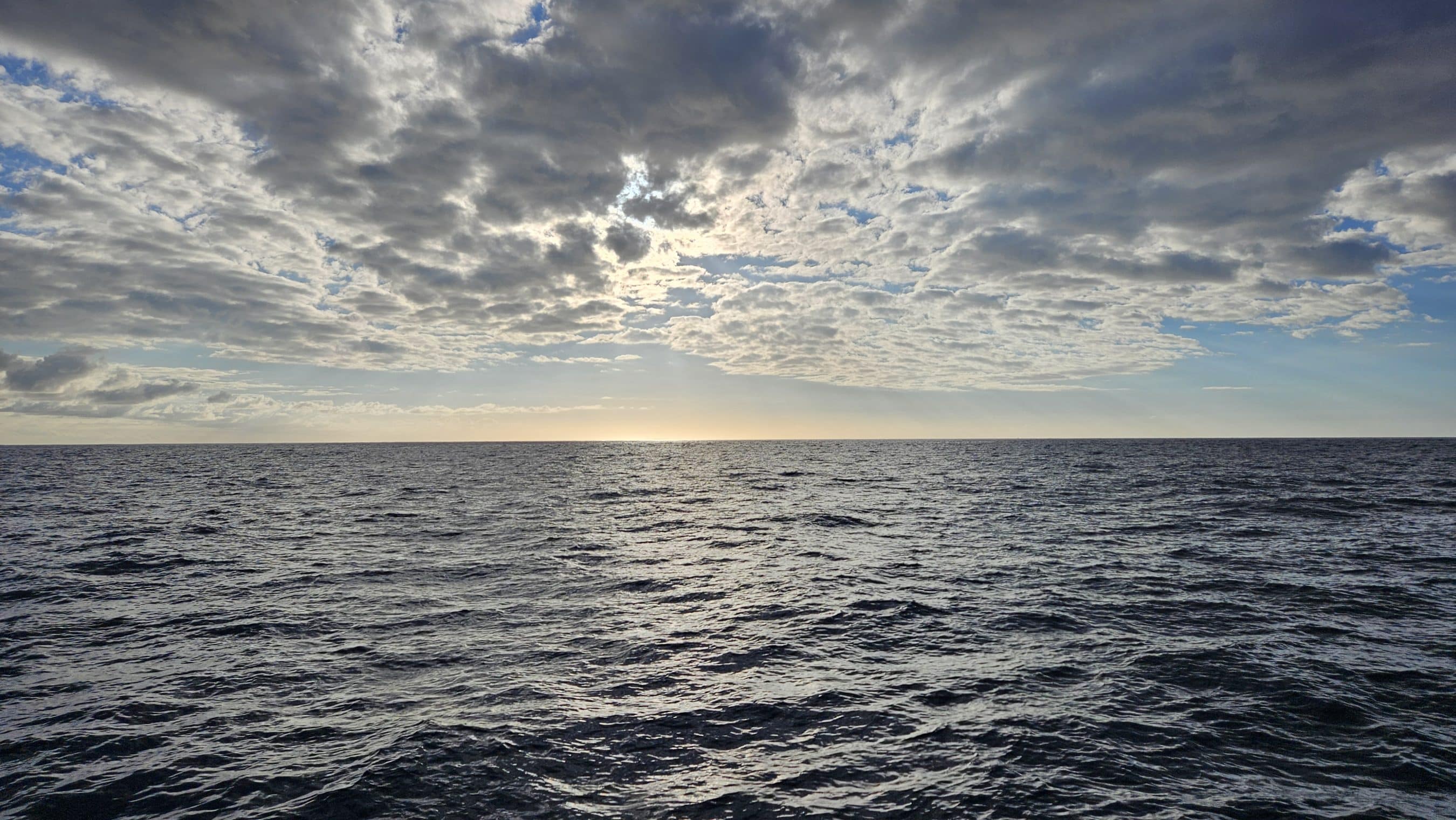 Ocean at sunset with a low horizon and cloudy sky; sunlight glints on the water.