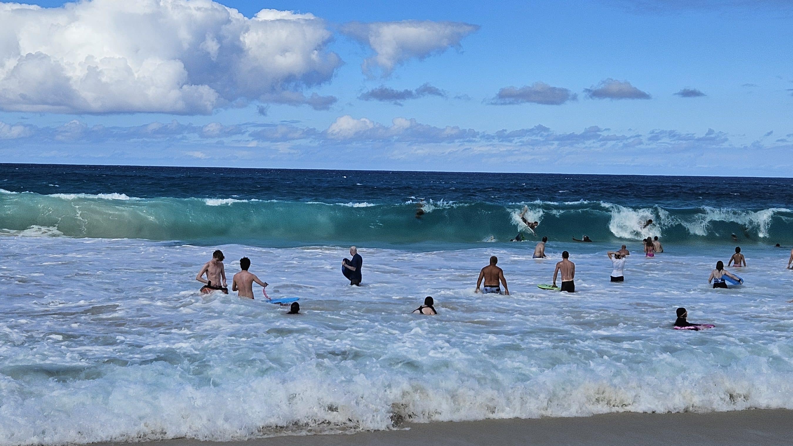 People enjoying a sunny beach, swimming and wading near rolling ocean waves.