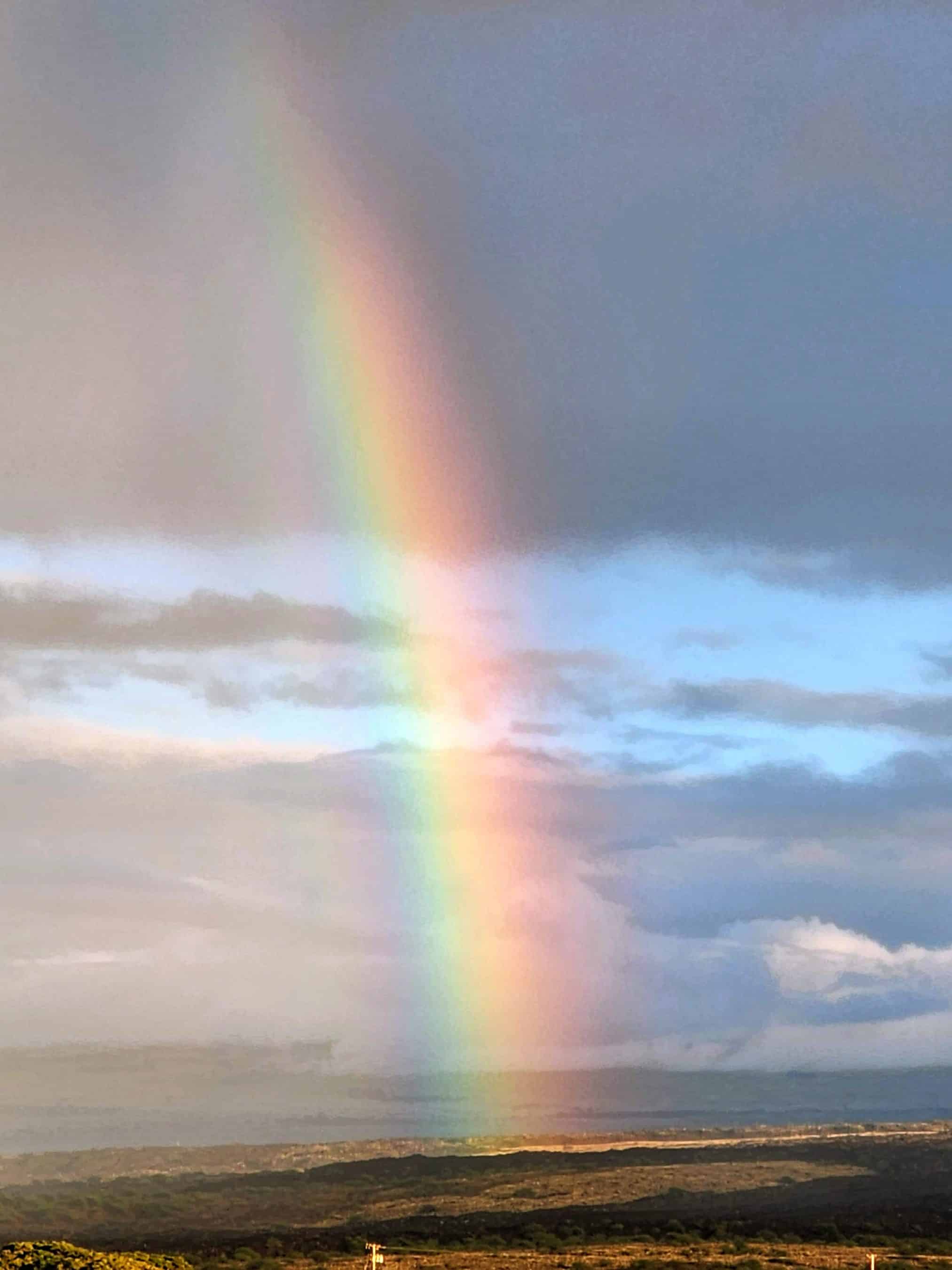 Rainbow arcing across a cloudy sky over a barren landscape with distant hills and a flat horizon.