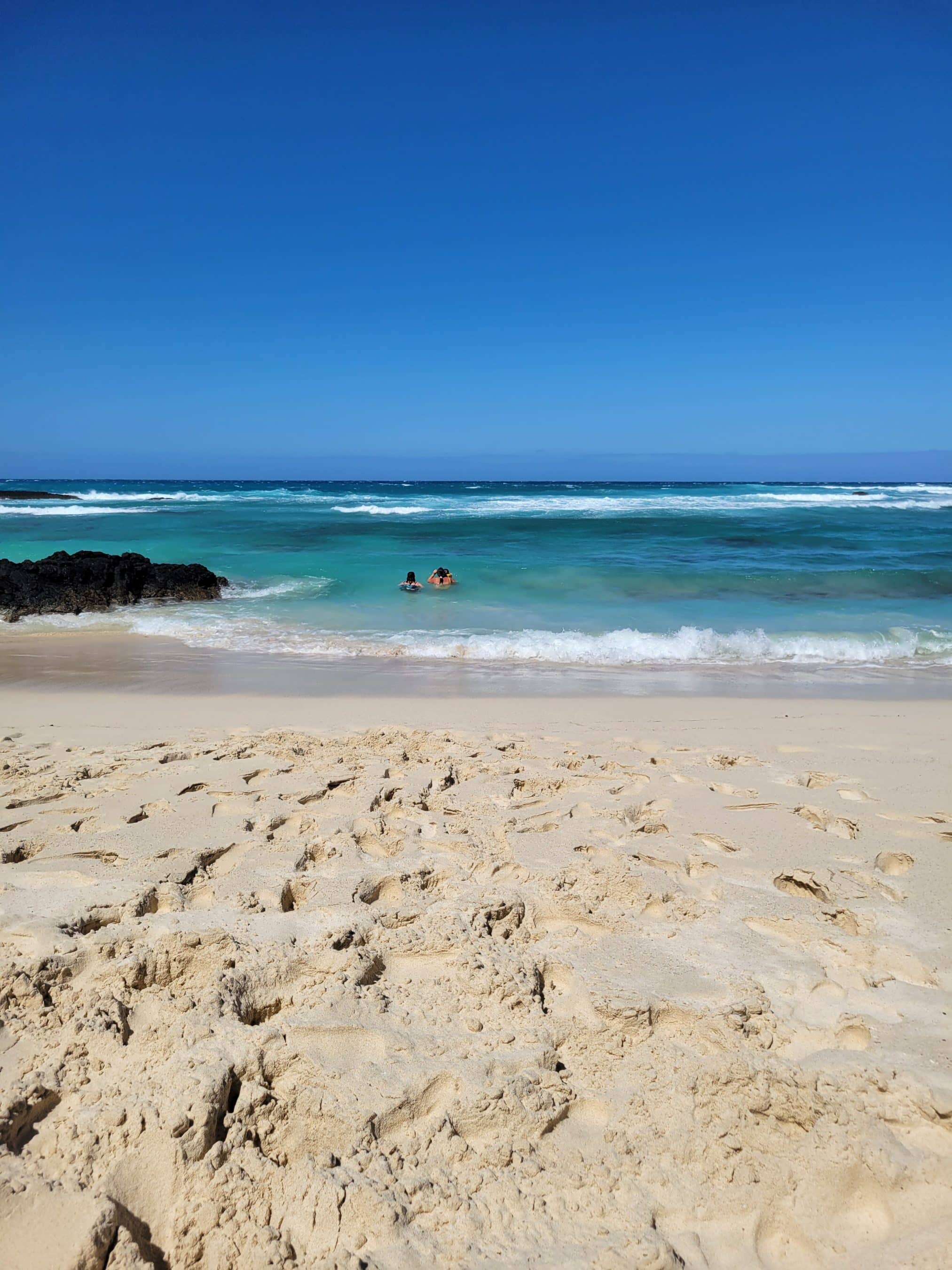 Sunny beach scene with turquoise water, white waves, and two swimmers near the shore; sandy foreground.