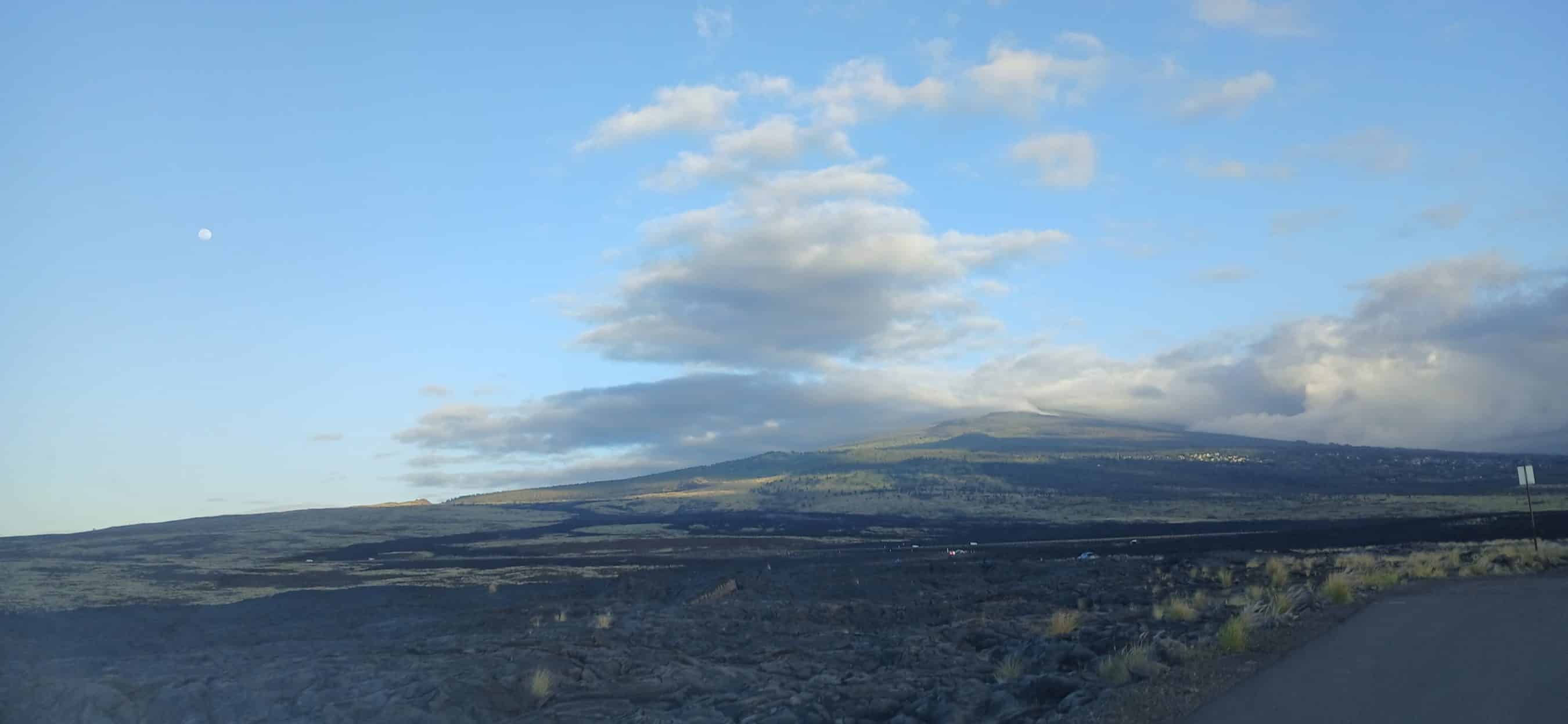 Wide landscape of a barren lava field and distant hills under a blue sky with scattered clouds and a small moon to the left.