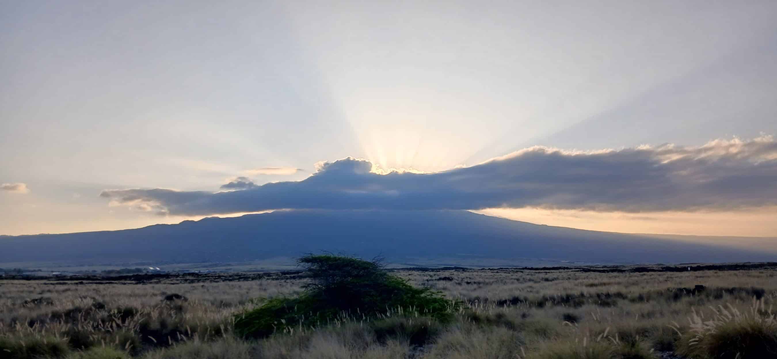 Sun rays stream from behind a low cloud over a flat grassland at sunset, with distant hills on the horizon.