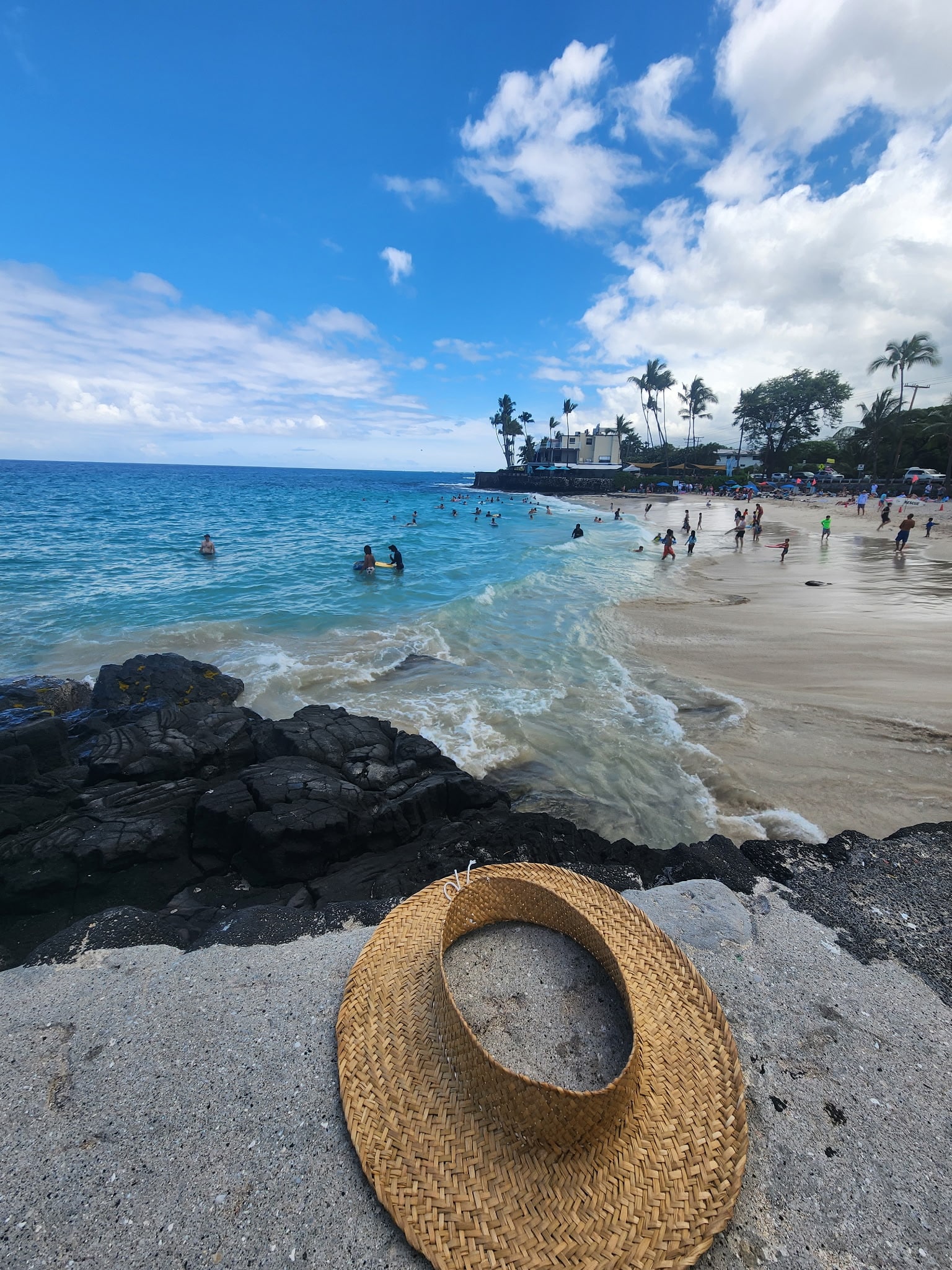 Small round straw hat rests on sandy beach with dark rocks in foreground, turquoise waves and people swimming in the water beyond.