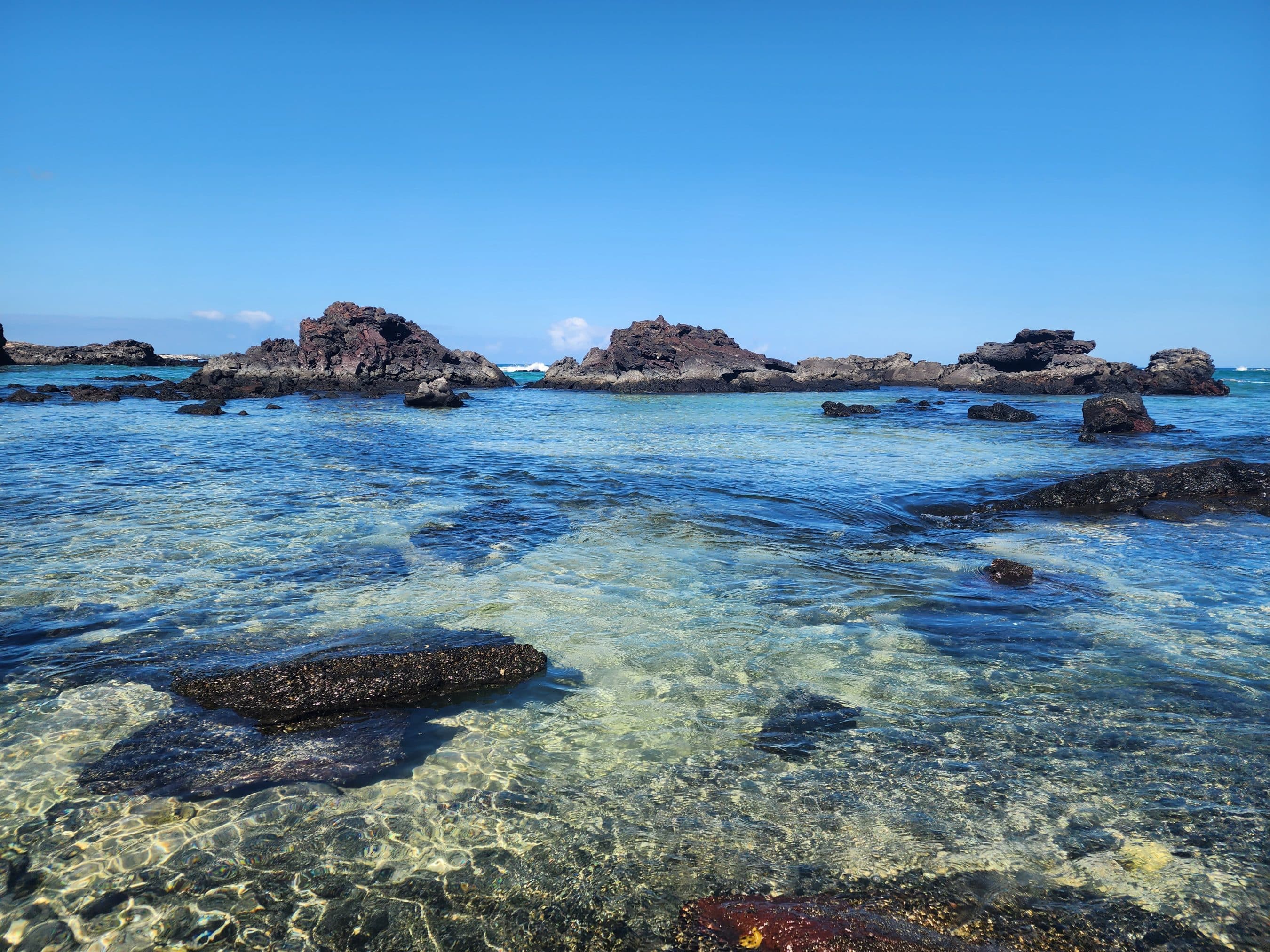 Clear turquoise sea with dark rocky outcrops along a shallow shoreline under a bright blue sky.
