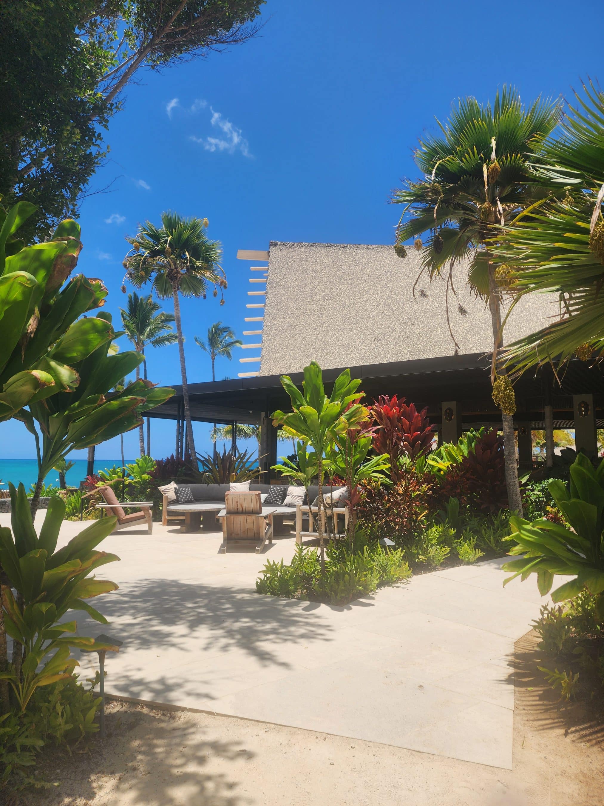 Beachside tropical patio with wicker chairs and a wooden table under palm trees, blue sky and ocean in the background.