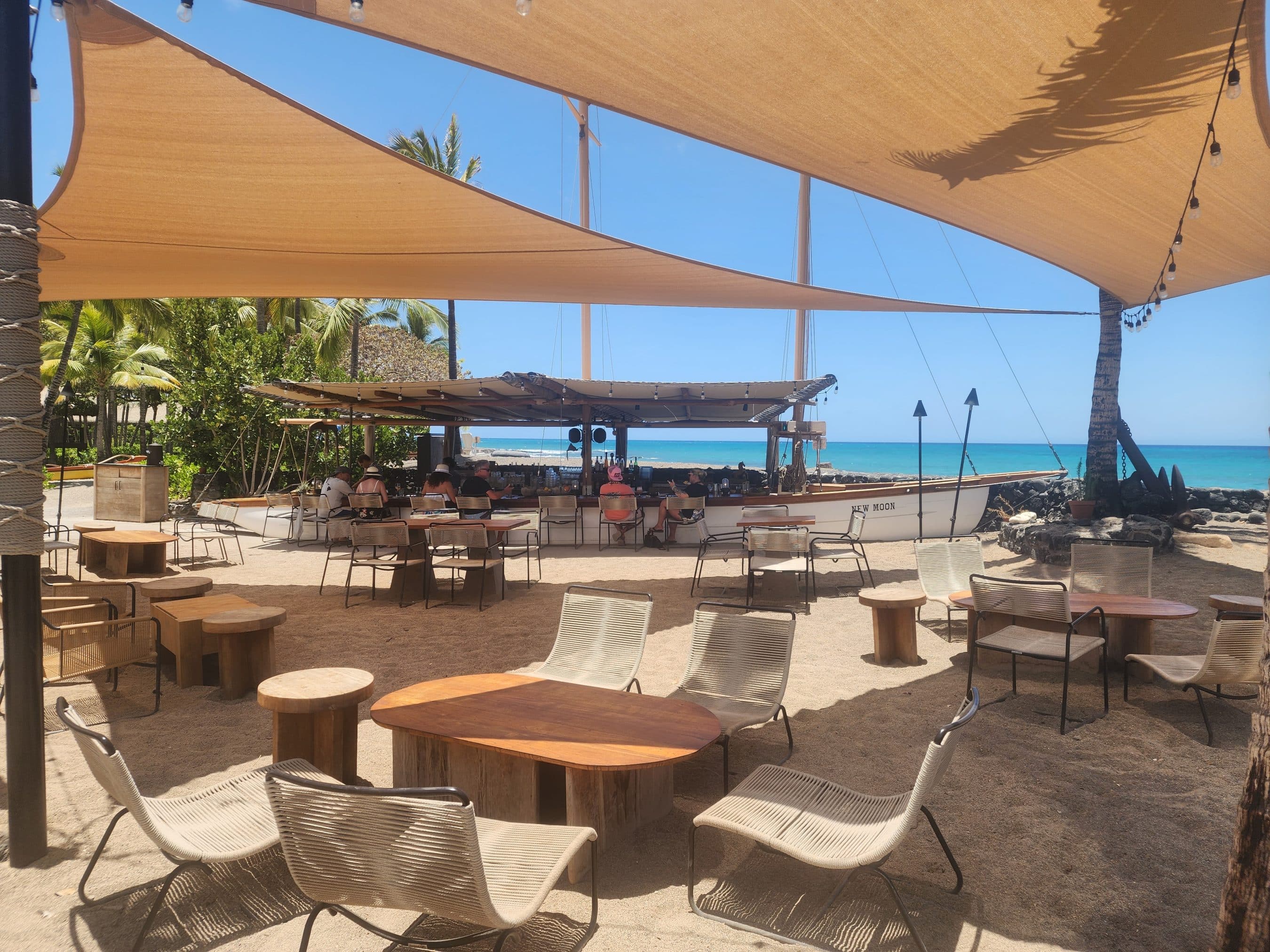 Outdoor beach cafe under beige shade sails with round wooden tables, wicker chairs, and the blue sea in the background.
