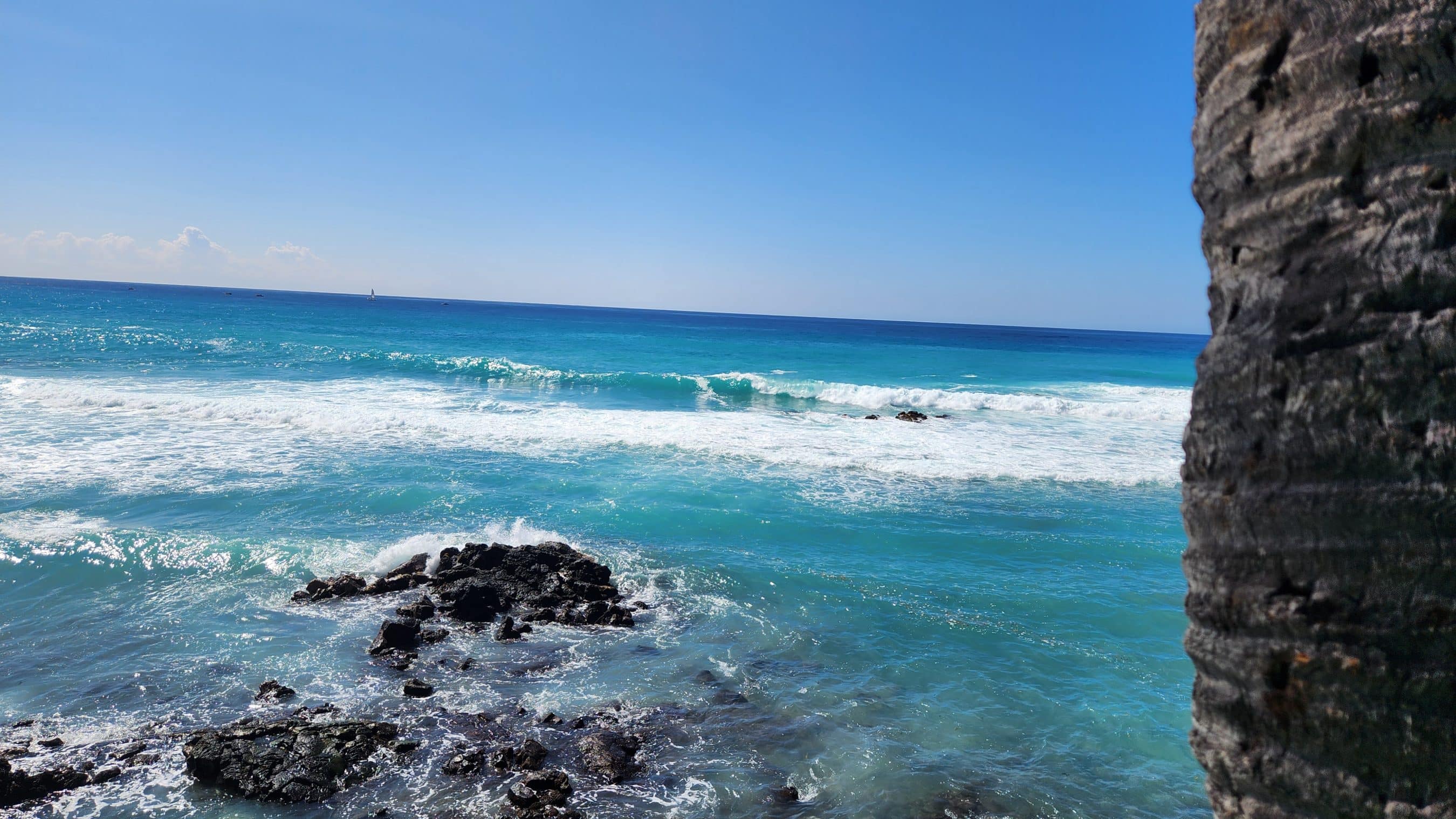 Turquoise ocean with white foam on rocks and a rocky cliff on the right under a clear blue sky