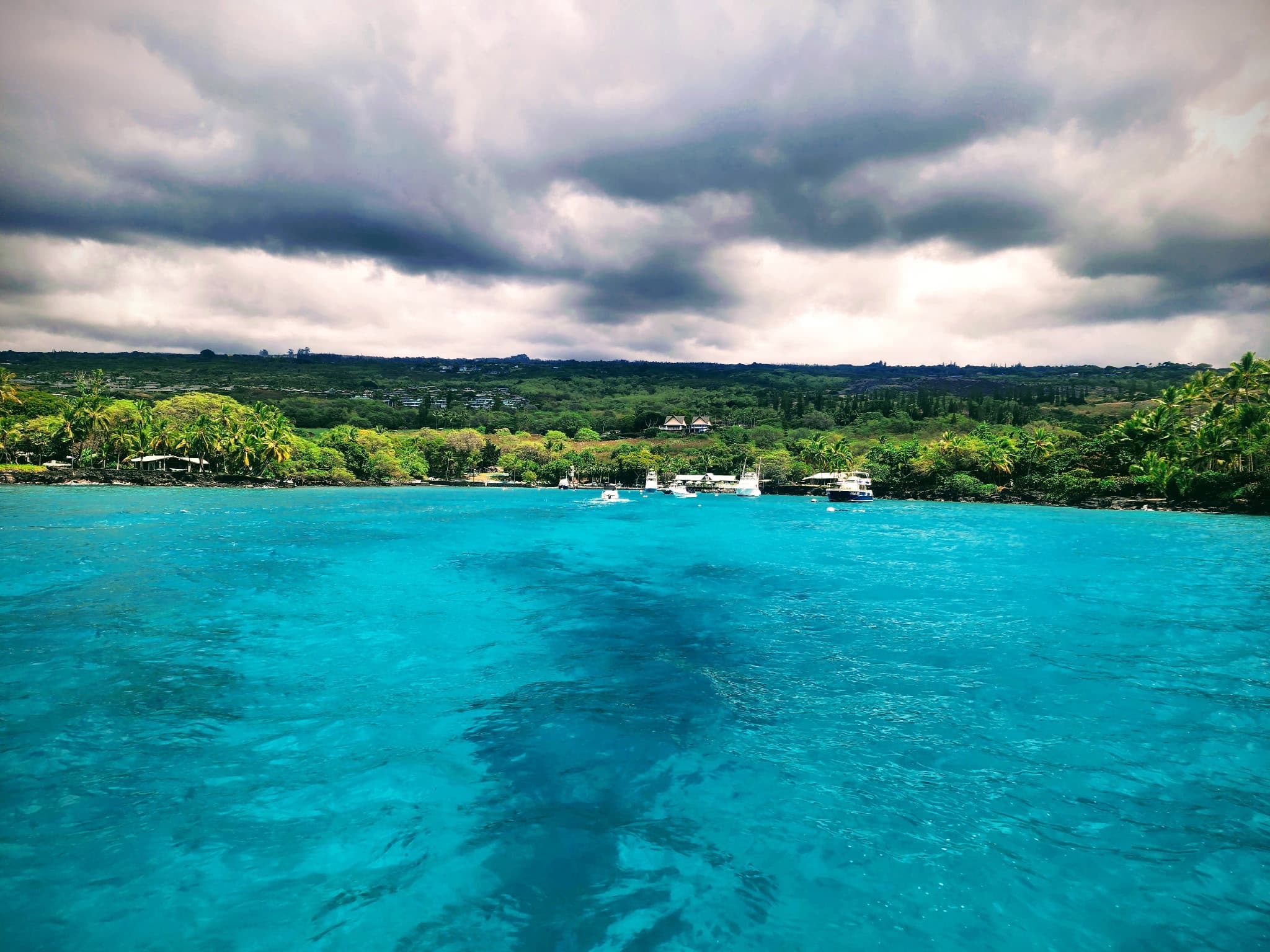 Stormy sky over a tropical shoreline with palm trees, green hills, and boats anchored along the turquoise sea.
