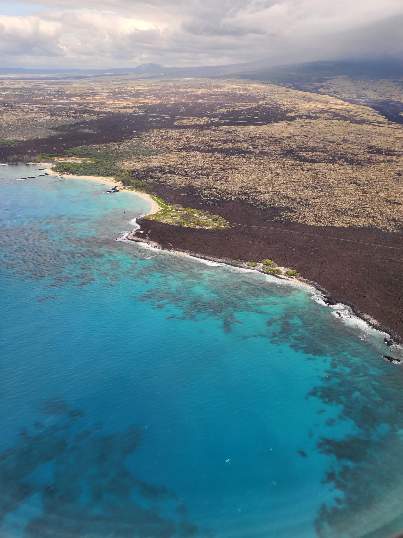 Aerial view of dark lava flows meeting bright turquoise ocean along a rocky coastline with a small sandy cove and greenery.
