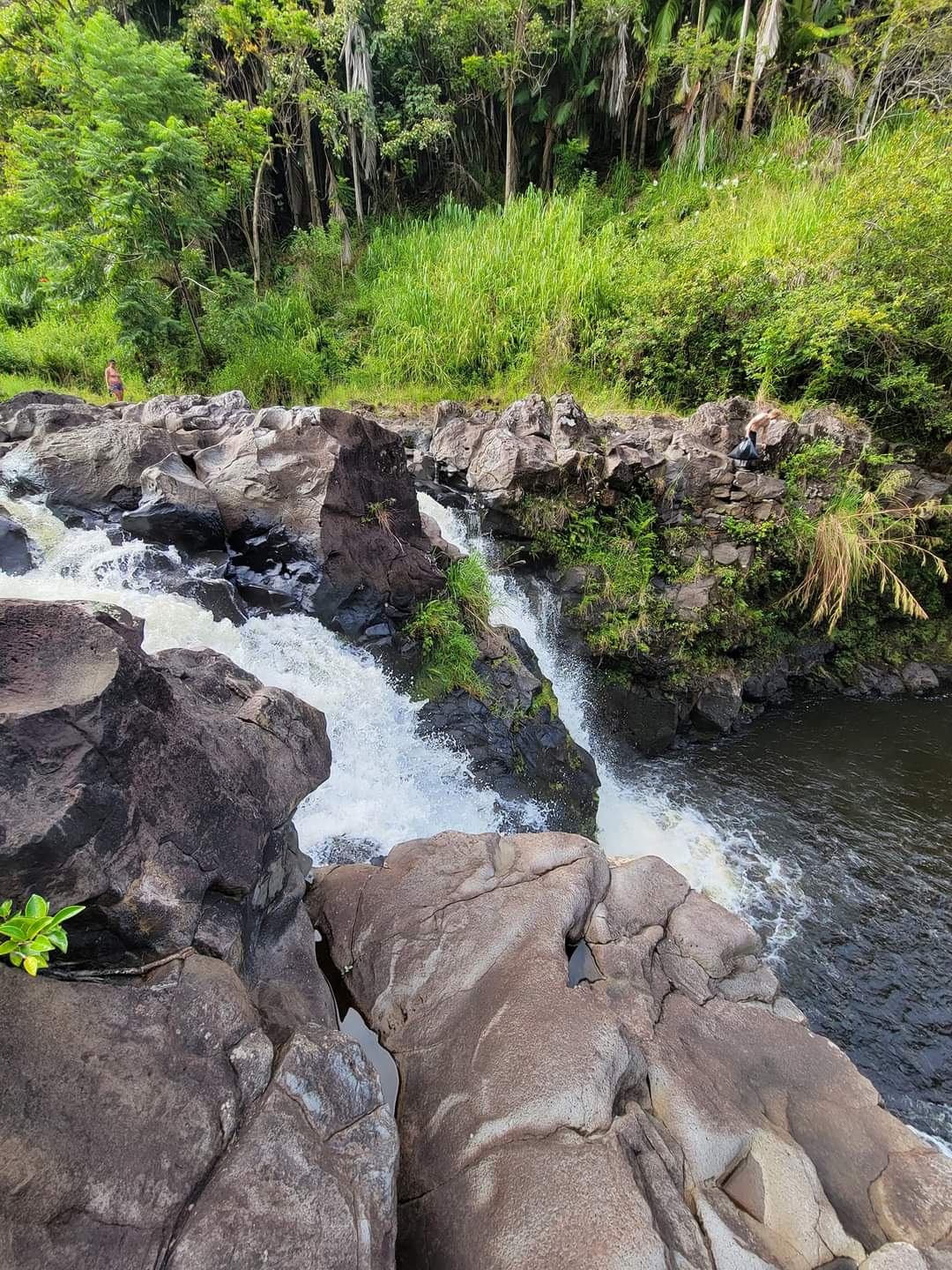 Rocky river with a small waterfall, lush green banks, and two people perched on the rocks.