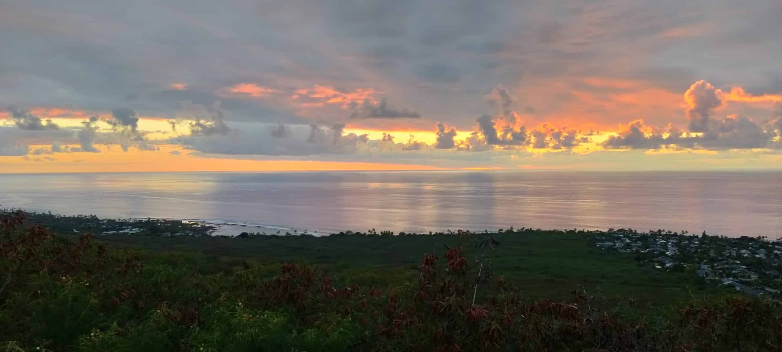 Sunset over a calm ocean with orange-pink clouds, green hills in the foreground, and a coastal town on the right.