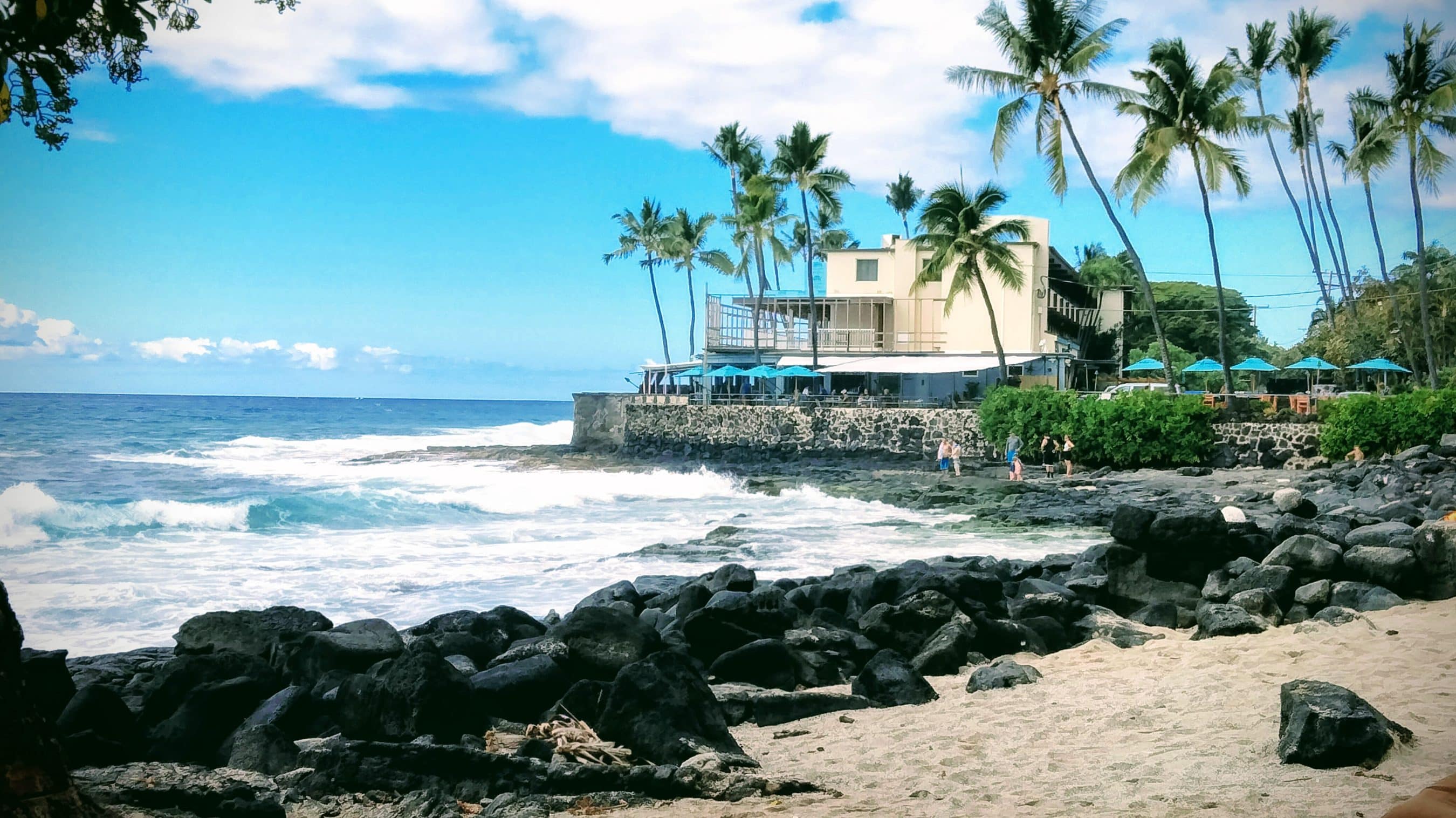 Beachfront restaurant and palm trees along a rocky shoreline with turquoise water and blue umbrellas under a bright sky.