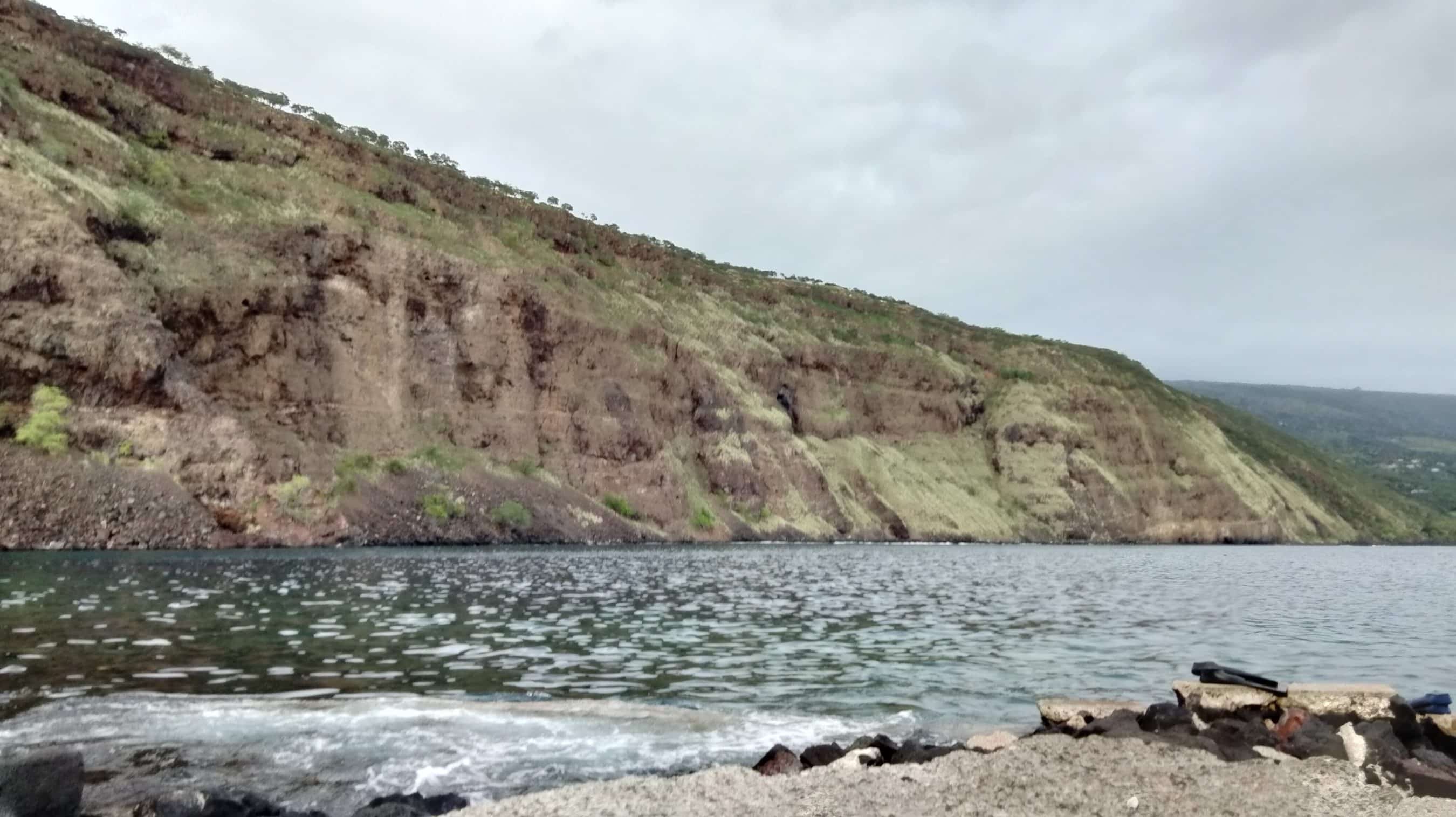 Steep rocky hillside along a calm body of water, with a pebbled shore in the foreground and a cloudy sky above.