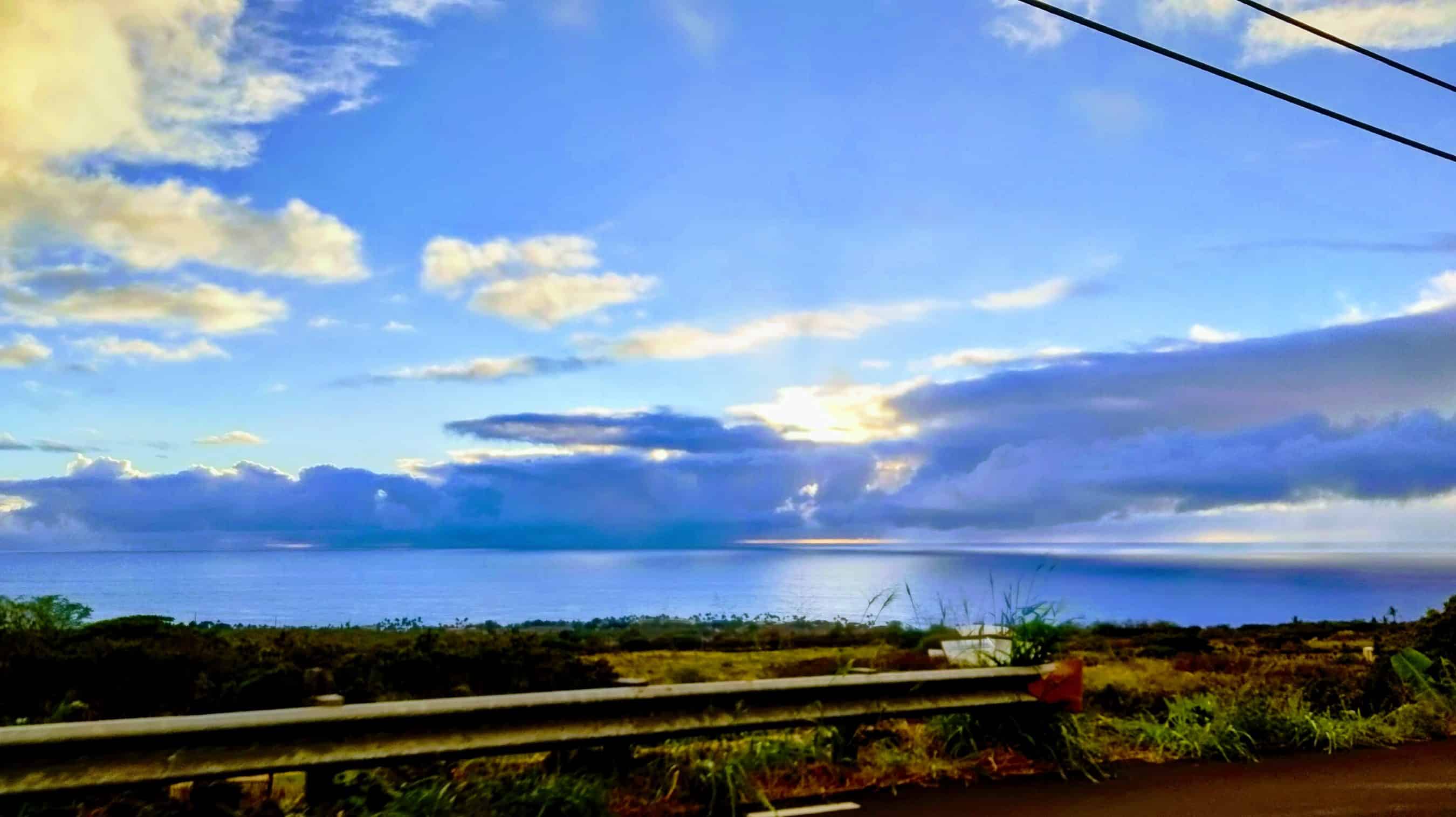 Coastal road scene with a guardrail in the foreground, calm blue ocean and cloudy sky above.