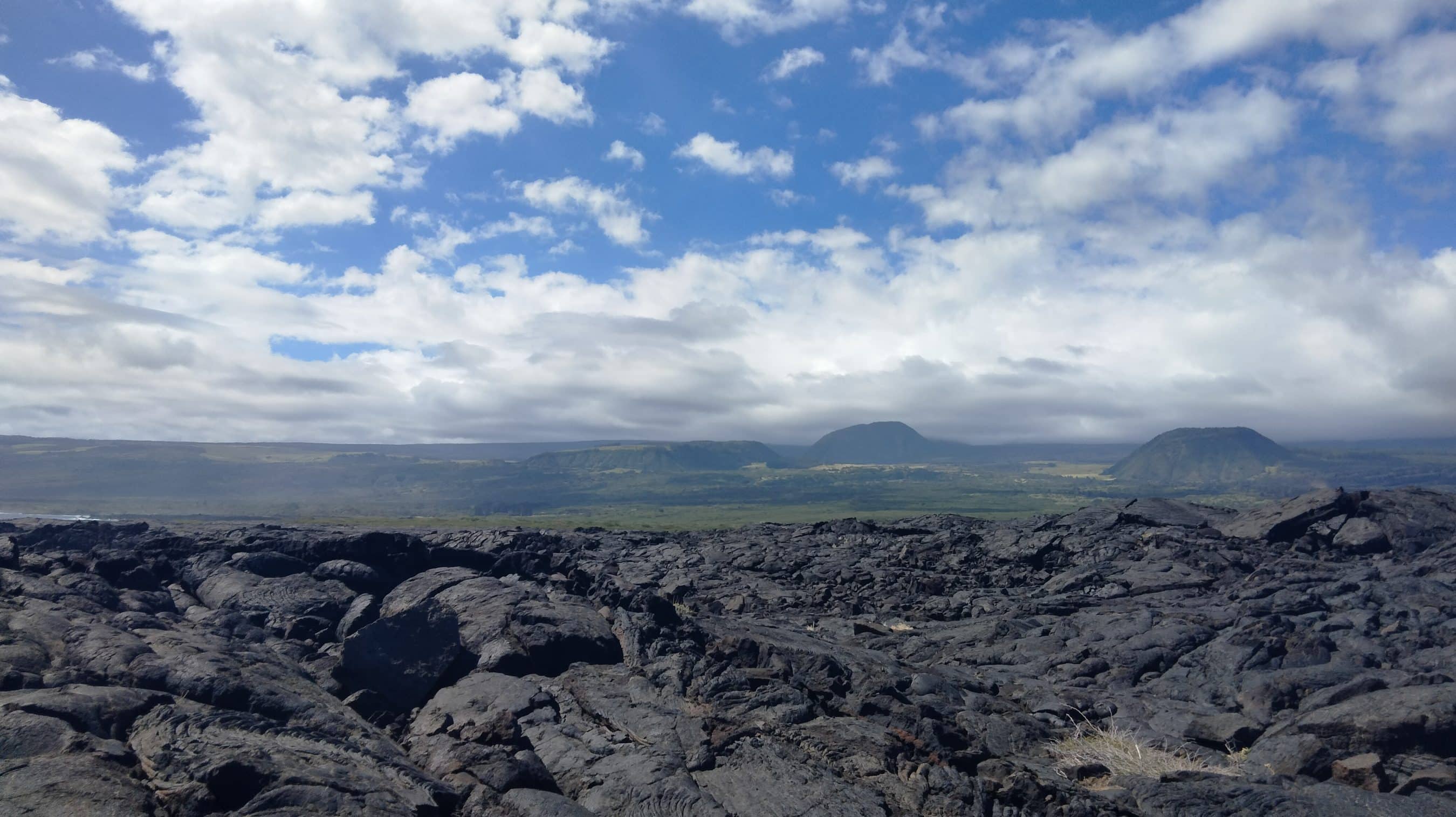 Dark, jagged lava rocks in the foreground with low hills on the horizon under a cloudy blue sky.