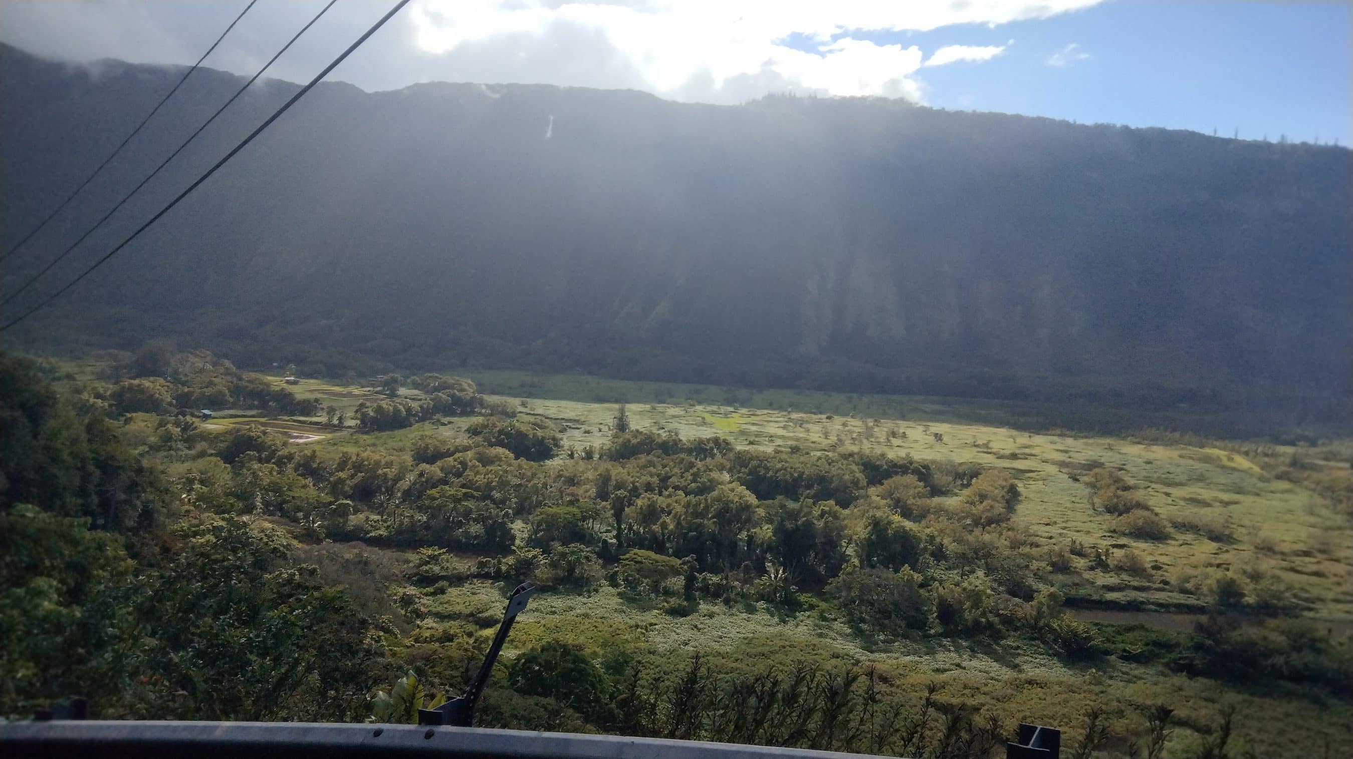Valley floor with fields and scattered trees, flanked by forested hills and a distant mountain ridge, with power lines crossing the view.