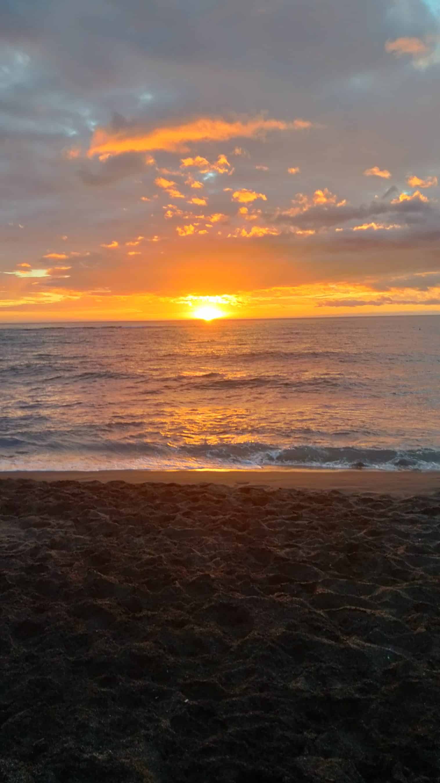 Sunset over the ocean with a golden glow on the water and a dark sandy beach in the foreground.