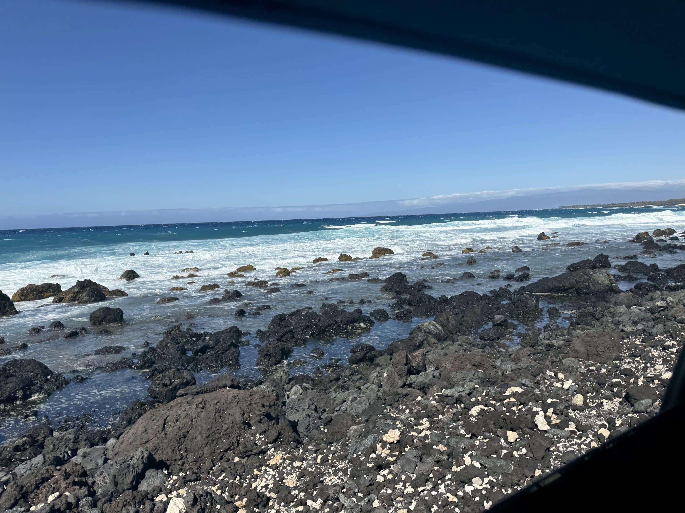 Rocky shoreline with dark volcanic rocks, turquoise water, and a clear blue sky. A sheltered viewing spot frames the top edge.