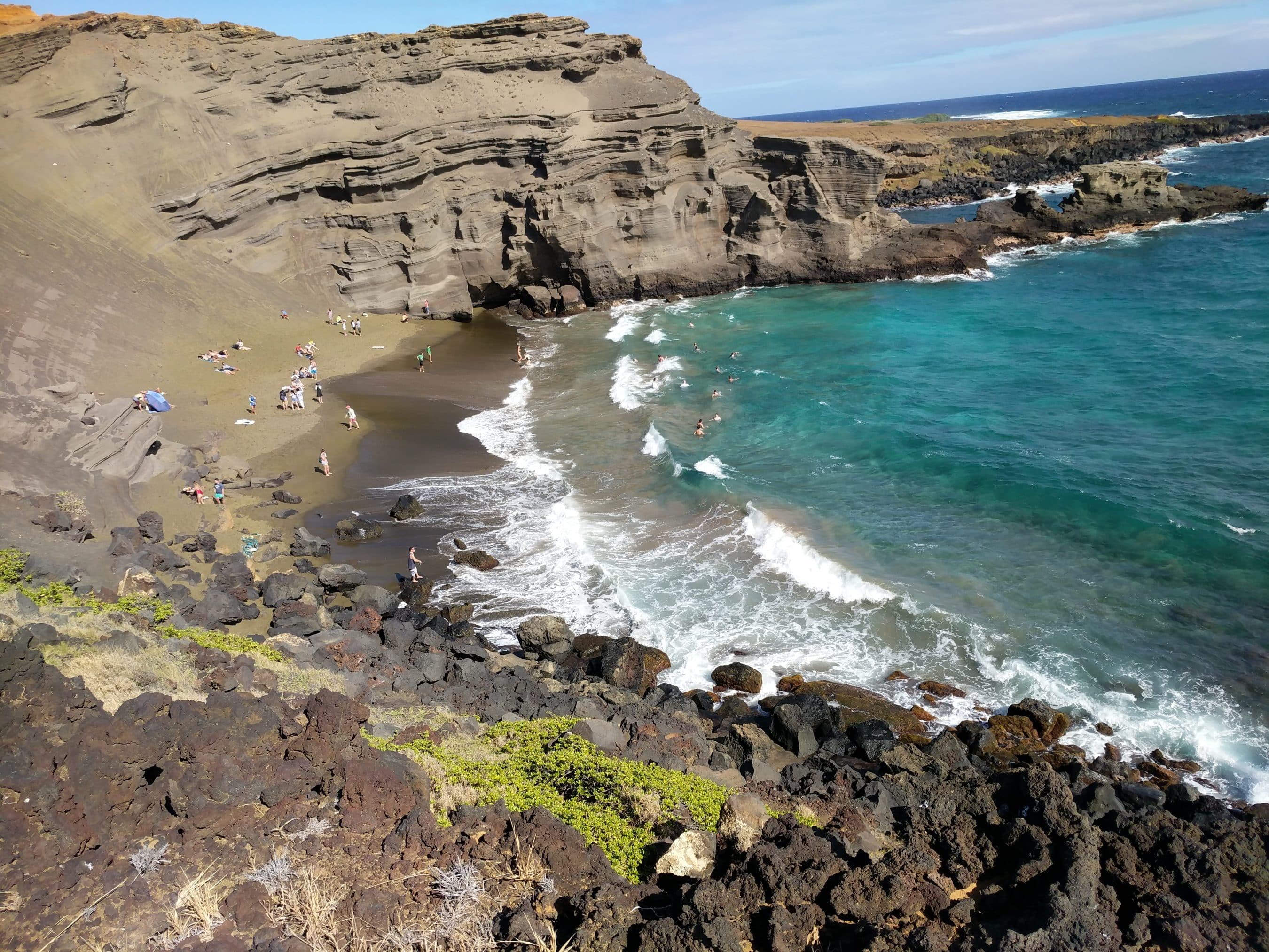 Cliffside coastline with layered rock formations, a sandy cove, and groups of people sunbathing and swimming in turquoise waters.