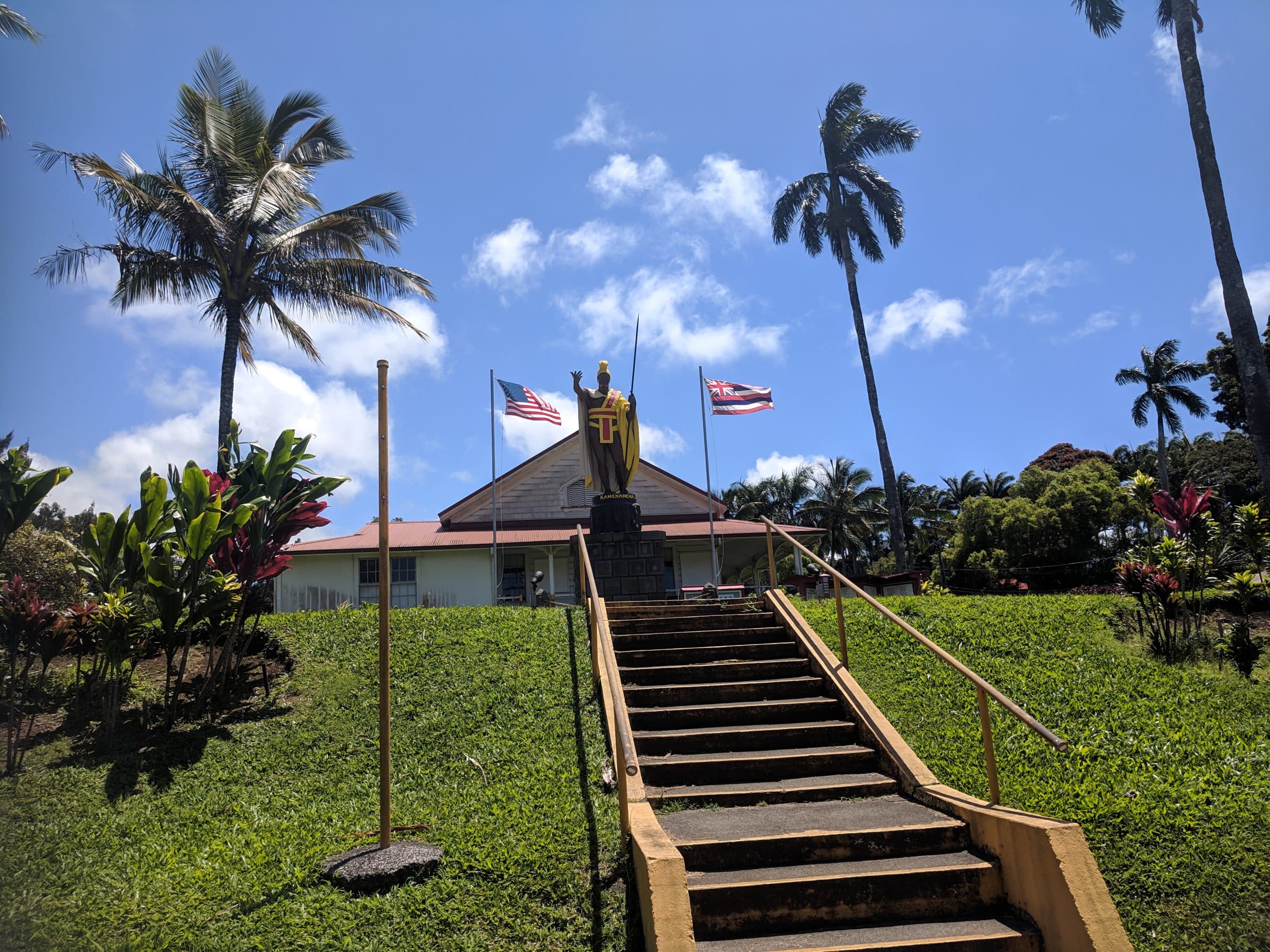 Steps and railing lead to a small building with a robed statue at the entrance, flanked by American and Hawaiian flags, under palm trees on a sunny day.