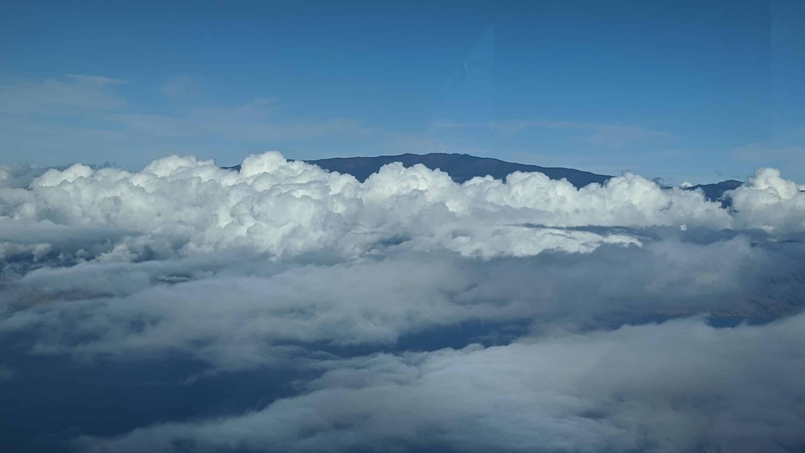 Aerial view of white fluffy clouds with a distant blue mountain range on the horizon under a clear blue sky.[shows horizon]”,