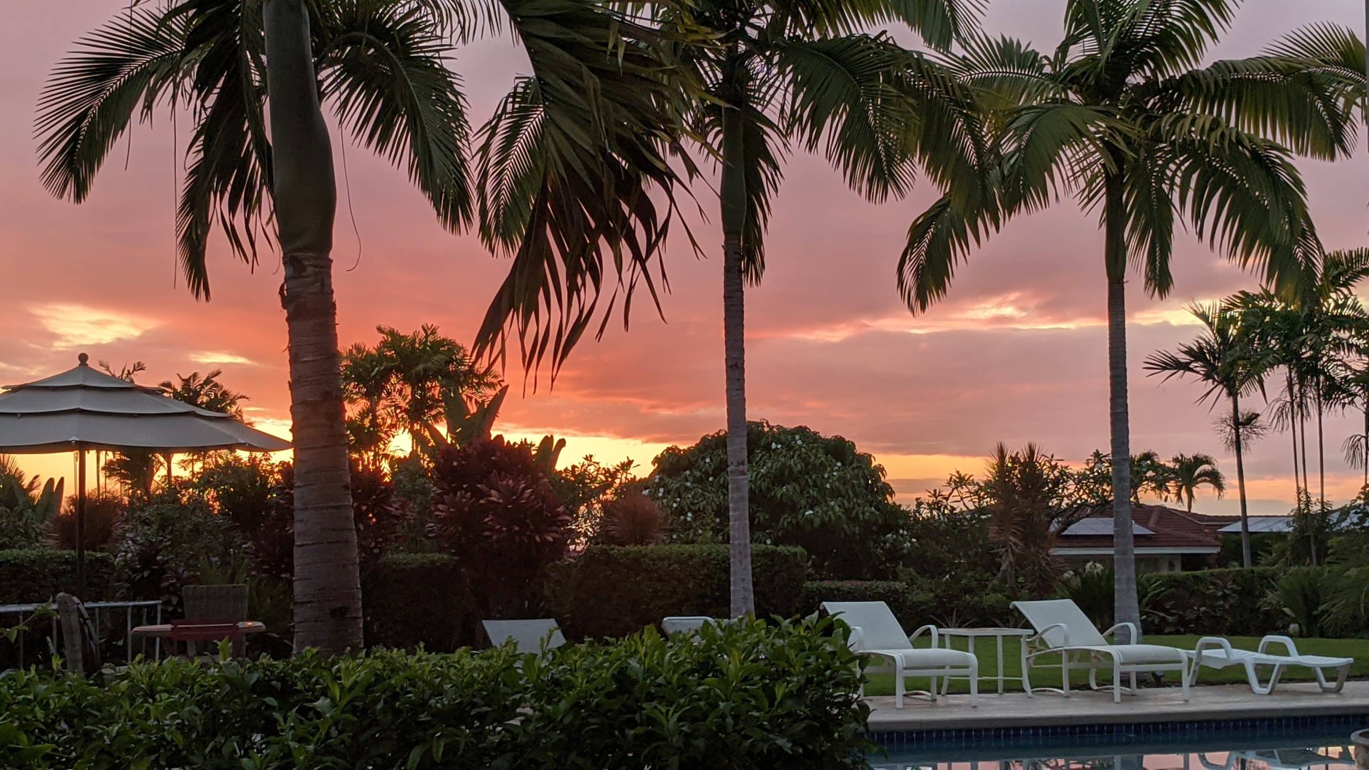 Sunset over a resort pool area with palm trees, lounge chairs, and umbrellas against a pink-orange sky.