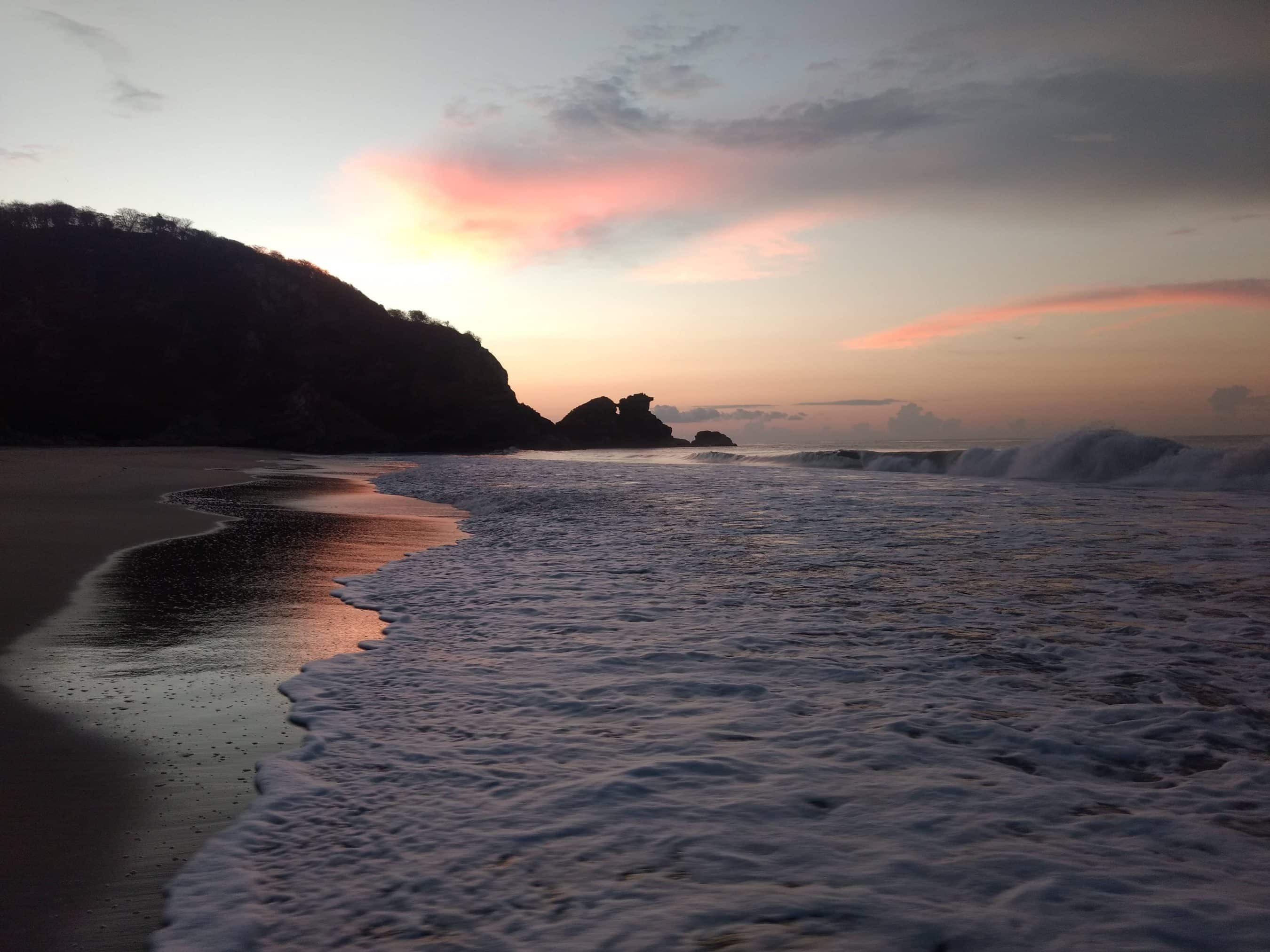 Sunset over a quiet beach with dark cliffs on the left and pink clouds reflecting on the water's edge.