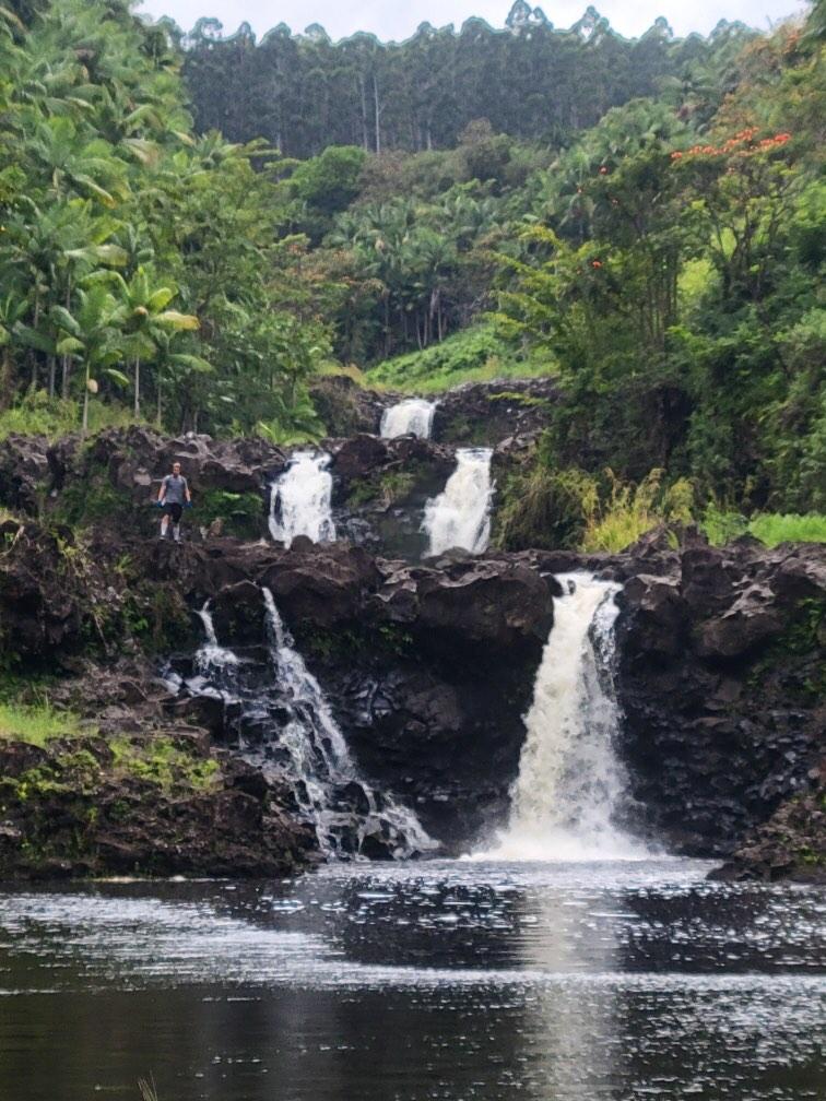 Waterfall flowing over dark rocks into a pool, surrounded by dense tropical vegetation, with a person standing on the left on the rocks.