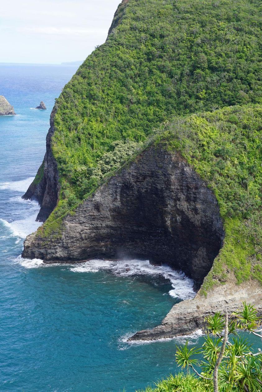 Steep green cliffside drops into blue ocean with a rocky shoreline and waves crashing at the base.