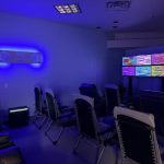 Dimly lit lounge with blue neon lighting, black leather lounge chairs arranged to face a wall of six colorful screens showing glitchy patterns.
