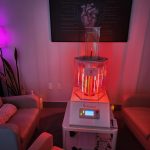 A purple-lit living room with a clear, multi-tube Biophoton device on a white table between two couches, pink glow reflecting on walls and a framed diagram hanging above.