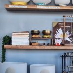 Wooden wall shelf display with meditation items: stacked books, brass sound bowls, a small Maneki-neko cat, a hanging jewelry stand, a potted trailing plant, and a framed lotus/flower poster against a blue wall.